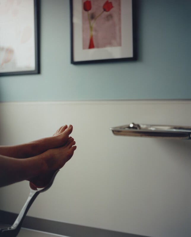 A pair of bare feet resting on a medical examination chair in a softly lit room. The wall has a light blue upper half and a white lower half. Two framed pictures of abstract flowers hang on the wall. A metal tray is mounted nearby.