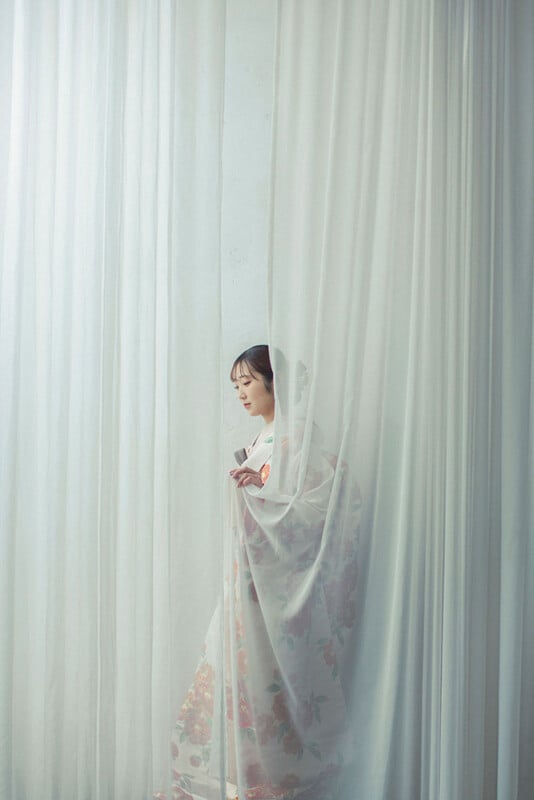 A person in a floral-patterned kimono stands behind semi-transparent white curtains, softly illuminated by natural light. They are looking downward, creating a serene and contemplative atmosphere.