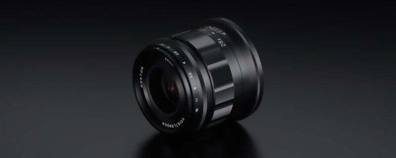 A black camera lens with various markings is positioned against a dark, reflective surface. The lens features focus and aperture adjustment rings, and the reflections highlight its metallic texture.