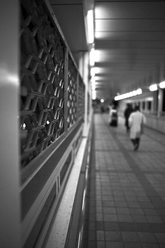 A black-and-white image of a long, tiled corridor with an intricate lattice design on the left wall. The corridor is lined with lights, and a person in a white coat is walking away in the distance.