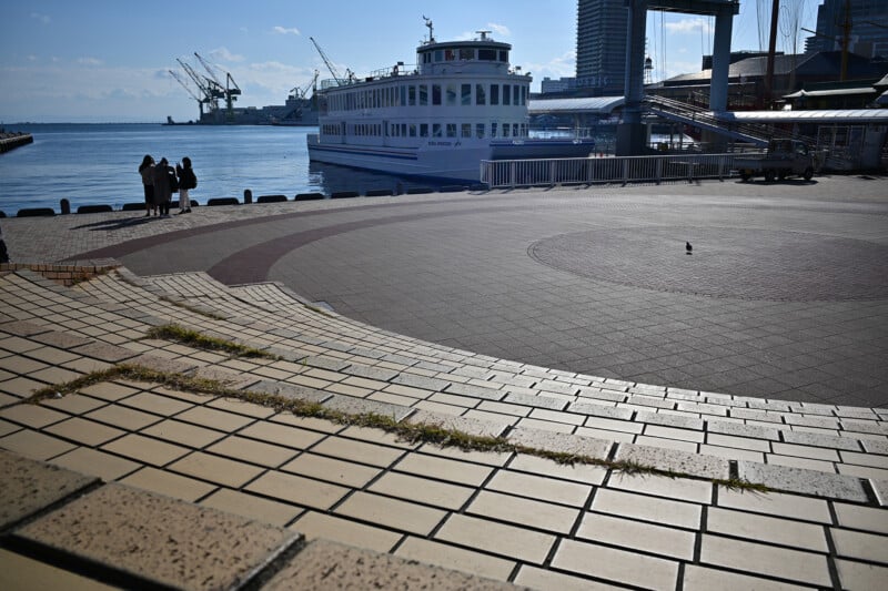 A waterfront scene with a large white ferry at the dock. Several people stand by the railing, overlooking the water. In the foreground, there are wide steps with grass growing between the tiles. Cranes and buildings are visible in the background.