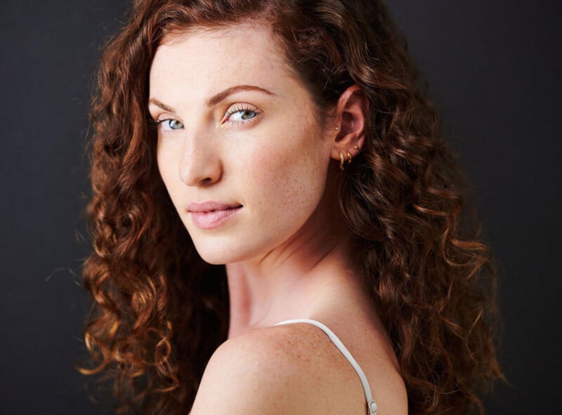 A woman with curly red hair and light eyes looks at the camera over her shoulder. She wears a white strap top and small gold hoop earrings, set against a dark background.