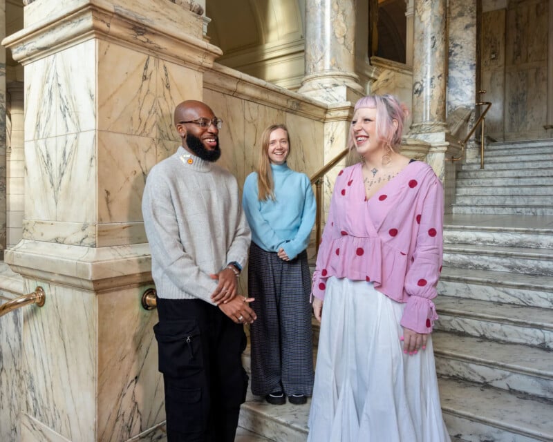 Three people stand in front of marble stairs in a grand building. The first person wears a gray sweater, the second person a light blue sweater, and the third person a pink blouse with a white skirt. They appear to be smiling and talking.