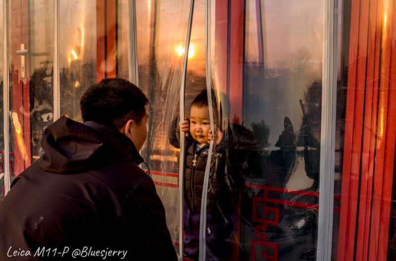 A child stands behind a clear plastic curtain, looking out with their hands on it, while an adult leans in from the outside. The sun casts a warm glow over the scene. Text in the bottom left corner reads, "Leica M11-P @Bluesjerry.