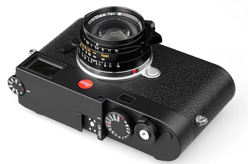 A black Leica camera with a textured body and a lens attached. The camera features dials and controls, including a red Leica logo on the front. The lens has distance markings in white and yellow. The view is angled from the top-right.