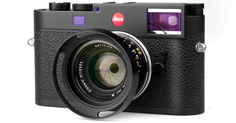 A black Leica camera with a textured body is shown with a large lens featuring a clear glass element. The camera has distinctive branding on the front, and various buttons and dials are visible on top.