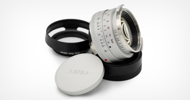 A silver camera lens with a visible focus and aperture ring, resting on a black lens cap labeled "Artra" and another cap in the background. The lens displays aperture markings from f/1.1 to f/16.