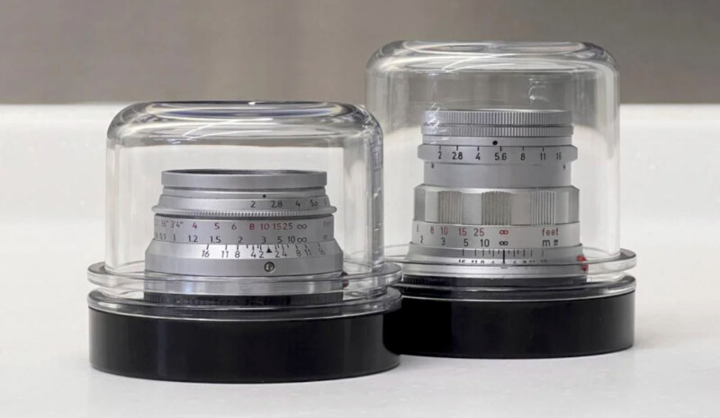 Two camera lenses encased in transparent glass domes on black bases. The lenses feature a metallic finish with engraved focus and aperture markings. They are displayed on a light surface with a blurred background.