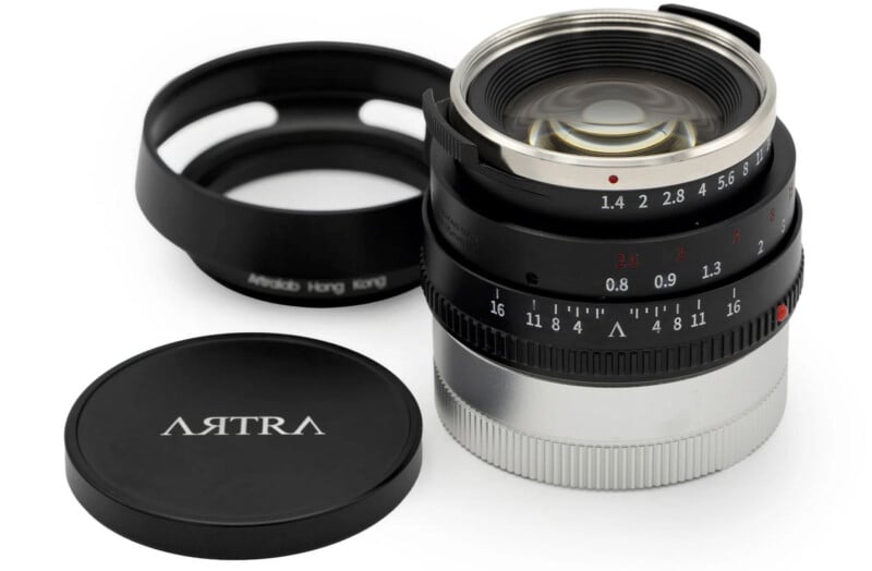 A close-up of a camera lens with a silver base and a black focus ring featuring aperture and distance markings. The lens cap is placed beside it, displaying the word "ARTRA." A hood is positioned in the background.