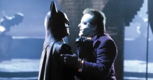 A man dressed as Batman stands face to face with another man dressed as the Joker. Batman is in a dark suit with pointed ears, and the Joker wears a purple suit with white face paint and a sinister expression. They are in a dimly lit setting.