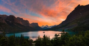 Stunning landscape of a tranquil lake surrounded by majestic mountains at sunset. The sky is a vibrant mix of orange, pink, and blue hues, reflecting on the water. A small island with trees is in the middle of the lake, framed by lush greenery.