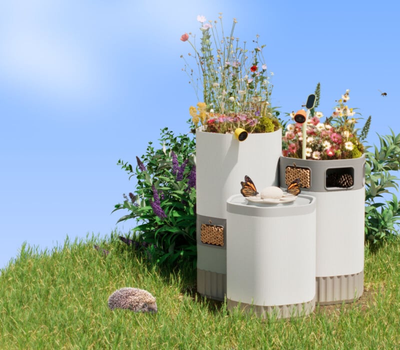 Three cylindrical planters with wildflowers and bee hotels are set on grass under a clear sky. A hedgehog is nearby, and butterflies and bees hover around the flowers, creating a vibrant and ecological garden scene.