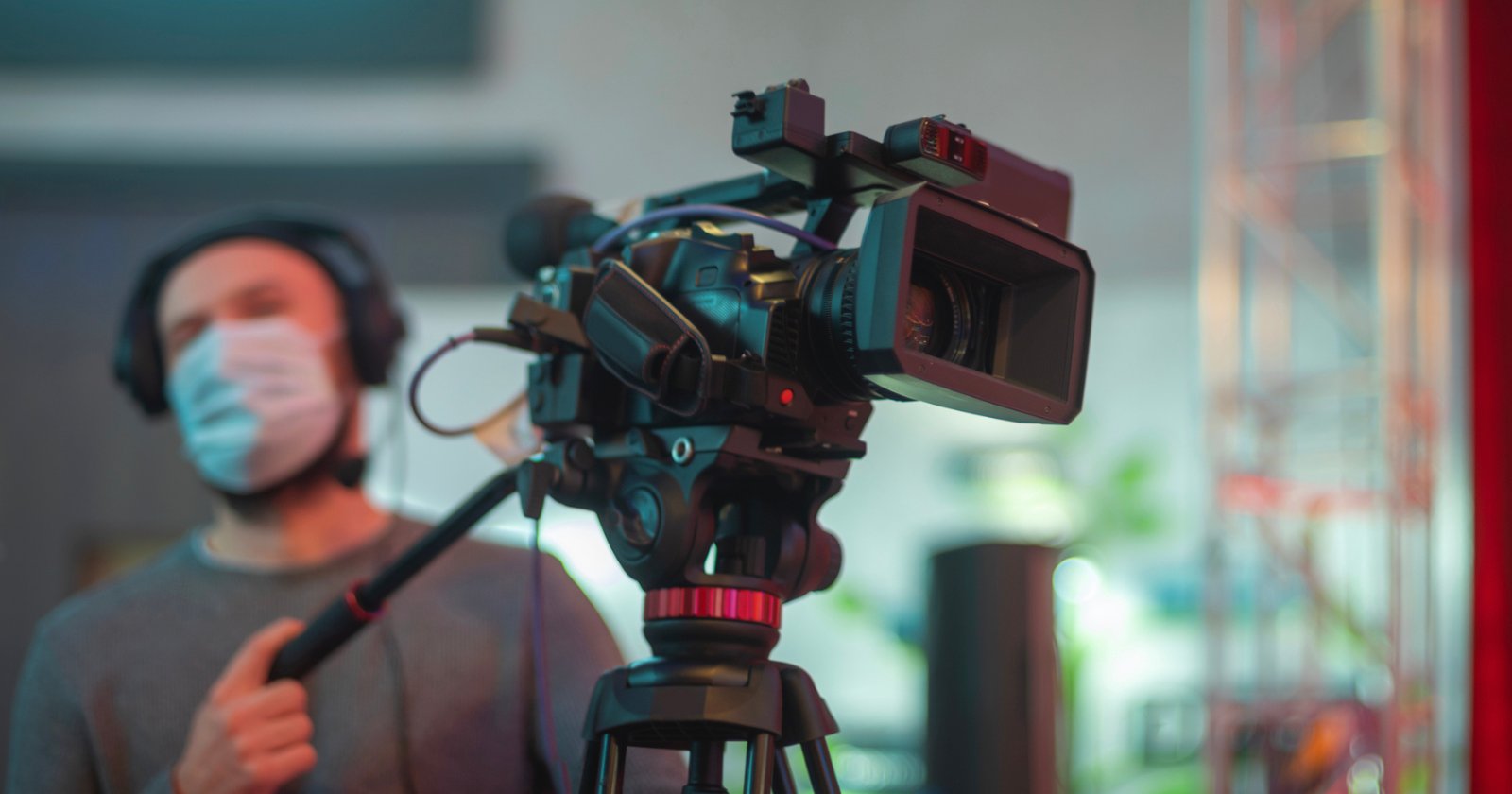 A professional video camera on a tripod is in focus, with a blurred background showing a person wearing a face mask and headphones, holding a microphone boom pole. The setting appears to be a studio or production environment.