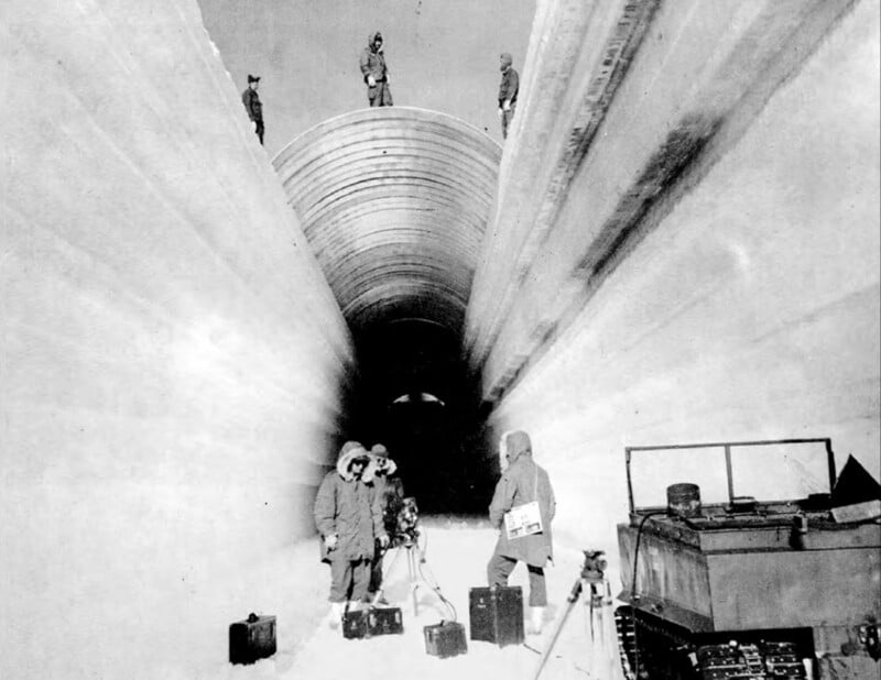 People in heavy winter clothing stand in a large, ice-cut tunnel. Various equipment is around them. Vehicles are visible to the right. Additional people are seen above the tunnel entrance.