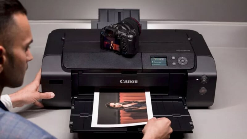 A person operates a Canon printer, printing a photo of a woman. A digital camera rests on top of the printer. The person adjusts the printer settings with one hand while observing the printed image.