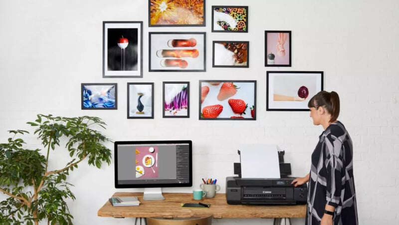 A person stands at a desk with a printer, feeding it a sheet. A computer monitor displays a photo editing program. Various framed colorful photos are arranged on the white brick wall above. A potted plant is on the left.