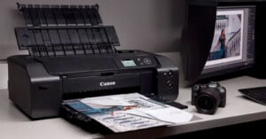 A Canon printer sits on a desk, printing a photo of a woman in a blue dress. Beside it, a DSLR camera and a monitor displaying the same image are visible. The setup appears to be in a photo editing or printing workspace.