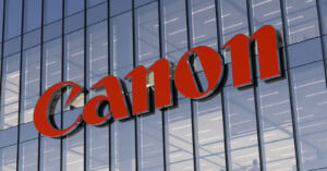 The image shows a glass building facade with a large red "Canon" logo prominently displayed. The reflective windows capture the sky and surrounding lights.