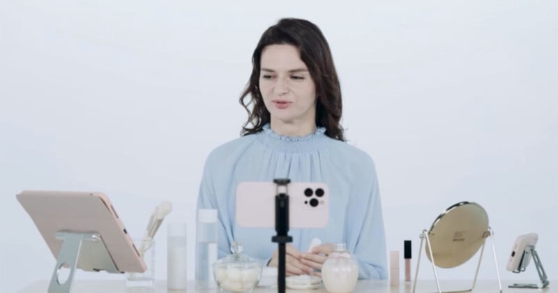 A person with long dark hair in a blue top is sitting at a table, facing a smartphone on a stand. The table has makeup products, a mirror, and a tablet. The background is plain white.