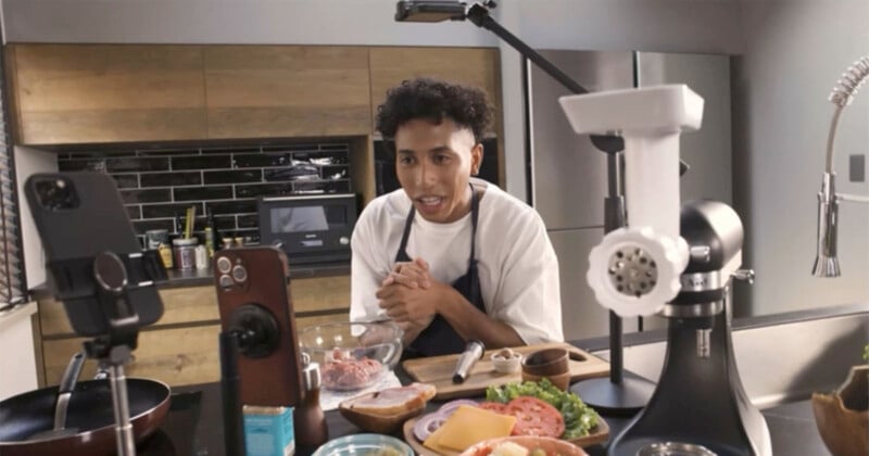 A person in a kitchen appears to be filming a cooking video. They are surrounded by ingredients like lettuce, tomatoes, cheese, and a burger patty, with cameras and a microphone set up in front of them.