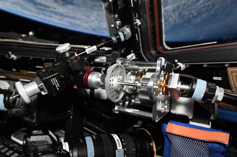 A close-up of a camera setup inside a spacecraft with Earth visible through the window. The equipment includes gears and attachments, highlighting advanced technology used in space exploration photography.