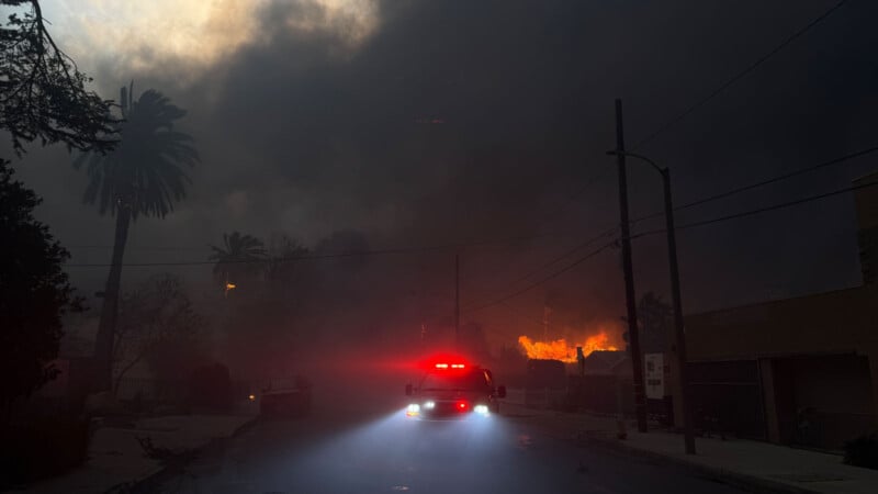 A fire truck with red lights illuminated drives through a smoke-filled street during a wildfire. Thick, dark smoke engulfs the sky, and flames are visible in the background. Tall palm trees and silhouettes of structures are partially visible.