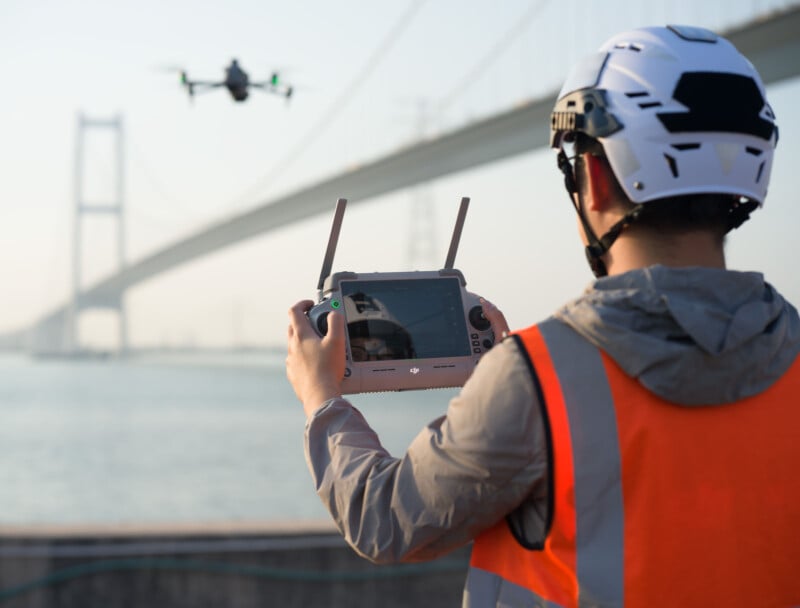 A person wearing a helmet and orange safety vest operates a drone using a remote control, standing by a large body of water. A bridge spans the background in a hazy sky.