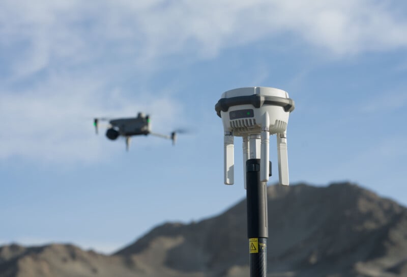 A drone flies in the sky over a rocky, mountainous landscape. In the foreground, a GPS device with antennas is mounted on a pole, with the clear blue sky stretching above.