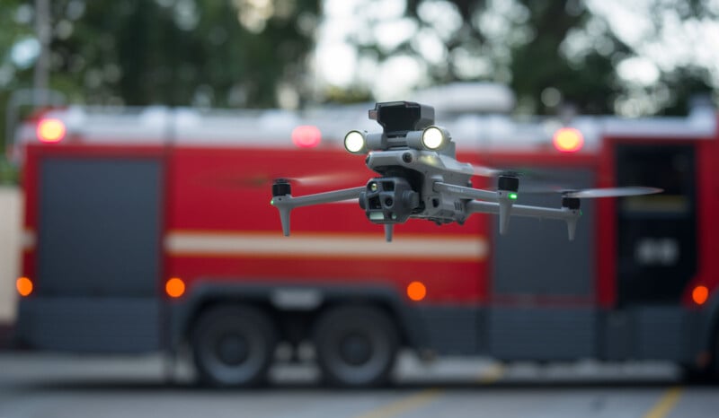 A drone with lights on is flying in front of a red fire truck. The background is slightly blurred, highlighting the drone in focus. The setting appears to be outdoors with trees partially visible.