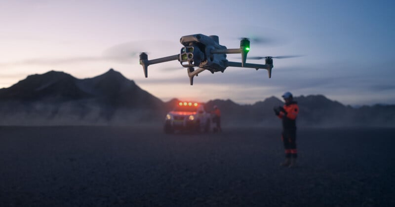 A drone hovers in the foreground at dusk, illuminated by green and red lights. In the background, a person in safety gear operates it, with a vehicle equipped with orange lights parked nearby. Mountains are silhouetted against the darkening sky.