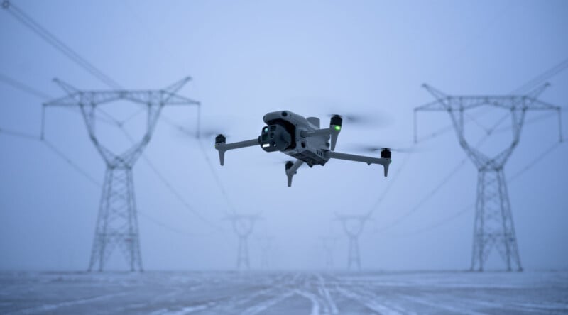 A drone hovers in the foreground with large power lines and towers in the background. The scene is set in a foggy, wintry landscape with snow covering the ground.