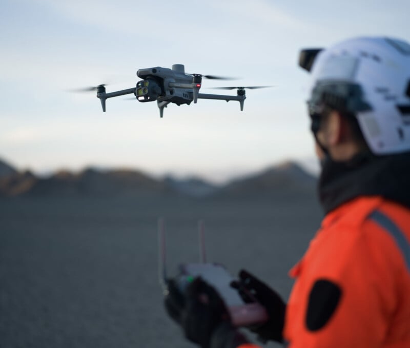 A person in an orange outfit and helmet operates a drone with a remote control. The drone is flying in the foreground over a rocky landscape with mountains in the background.