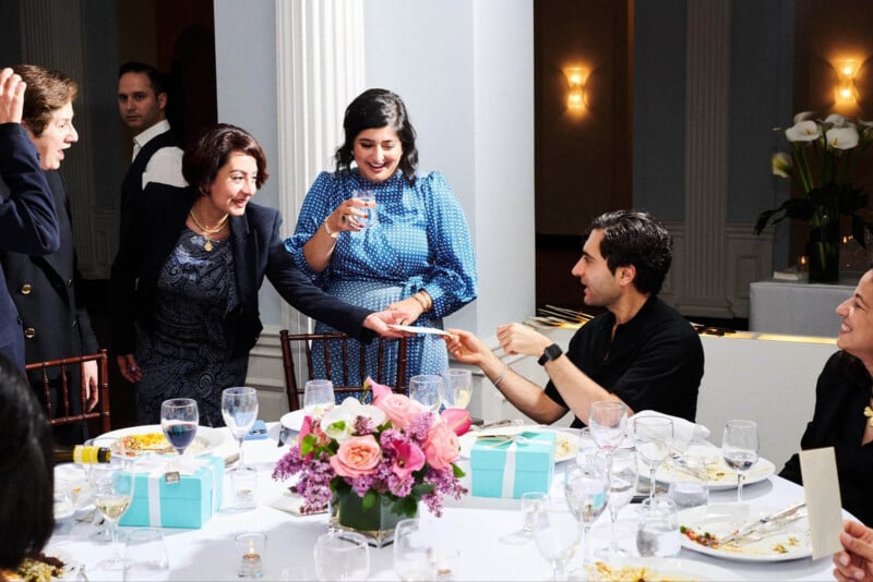 A group of people seated around a table at a formal event. One person hands a card to another. The table is decorated with a floral centerpiece and Tiffany-blue gift boxes. Plates with food and glasses are on the table. People are smiling and interacting.