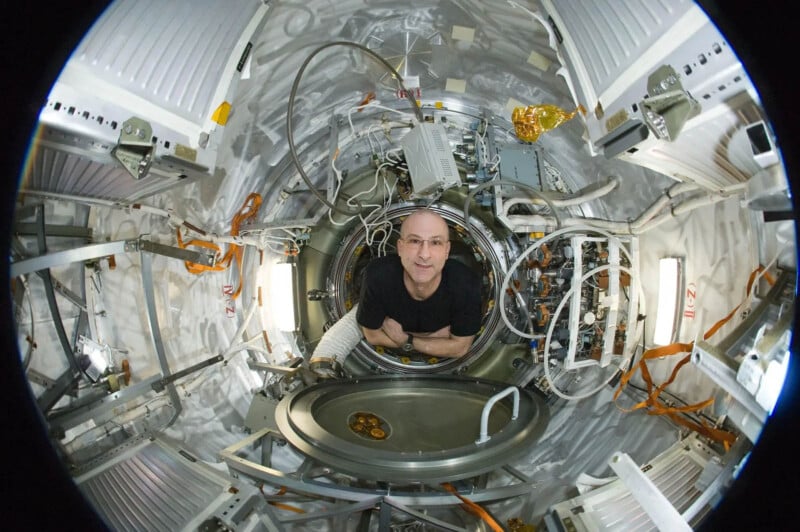 A person floats inside a space module, surrounded by cables and equipment. The interior is lined with metallic panels and orange straps, with a round hatch visible. The image has a fisheye lens effect, creating a wide, curved perspective.