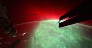 A view of the Earth's horizon from space, featuring a vivid display of red and green auroras. Parts of a space station, including solar panels, are visible against the backdrop of the glowing atmosphere.