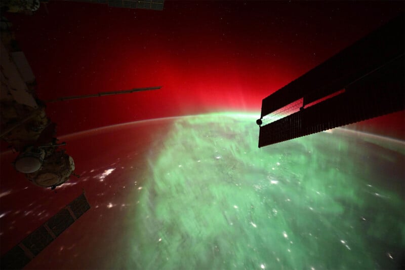 View from space showing vibrant green and red auroras over Earth. Parts of a spacecraft are visible, with solar panels on the right. Stars dot the dark sky, creating a striking contrast with the colorful auroras below.