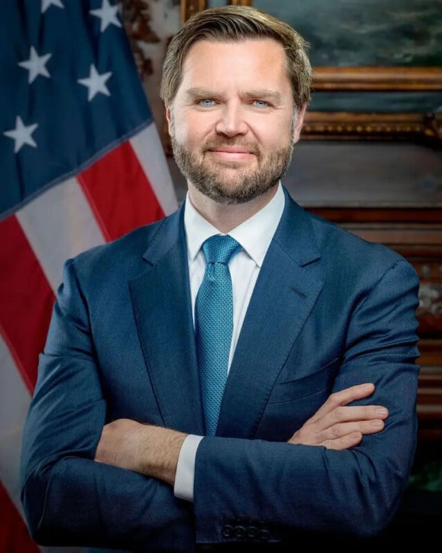 A man dressed in a blue suit and tie with arms crossed stands smiling before a U.S. flag. The background features a framed painting and elaborate wood features.