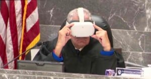 A judge wearing a virtual reality headset sits at a desk in a courtroom. An American flag is visible in the background, and a computer monitor is on the desk.