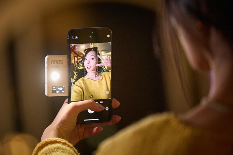 A person in a yellow sweater holds a smartphone to take a selfie, with an attached light illuminating their face. The background is softly blurred, emphasizing the device and the person on the screen.