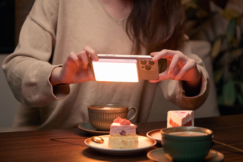 A person holds a smartphone horizontally over two slices of cake on plates and two cups of coffee on a wooden table. The phone emits a bright light, illuminating the treats, creating a cozy atmosphere.