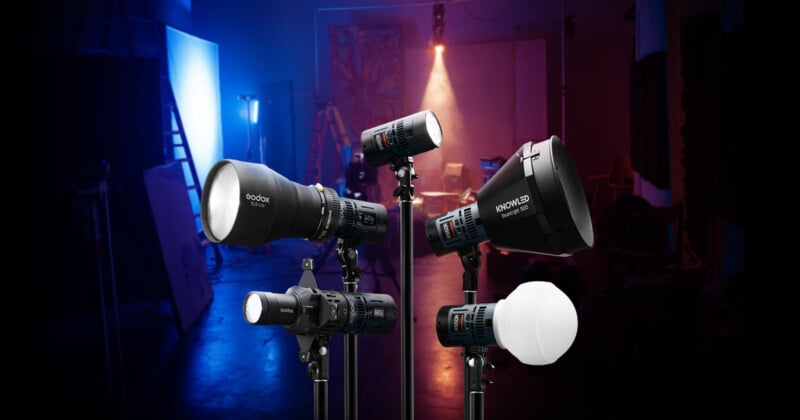 A variety of studio lights on stands are showcased in a dimly lit room with colored lighting in the background. The equipment includes different models and shapes, designed for professional photography or video production.