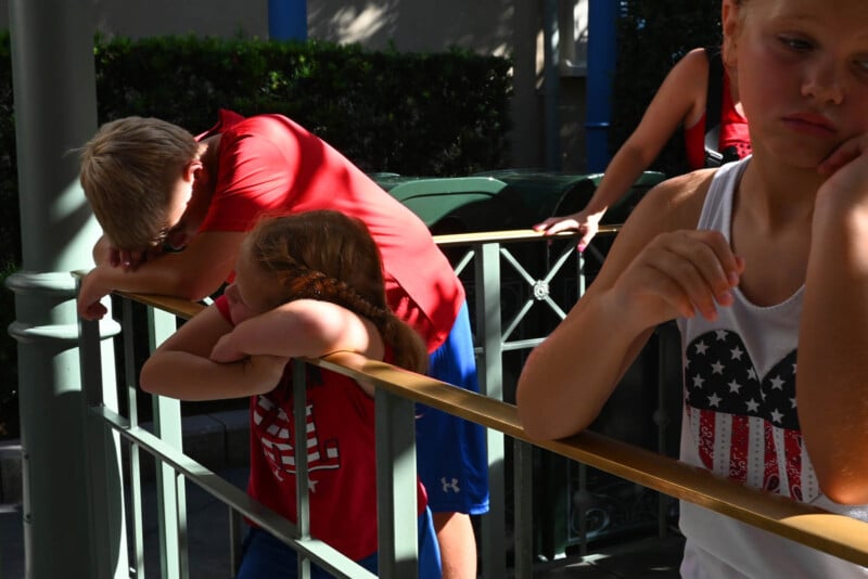 A group of children wearing red and white American flag-themed shirts lean on a metal railing. They appear tired or bored, with one child resting their head on their arms. The background features greenery and more railings.