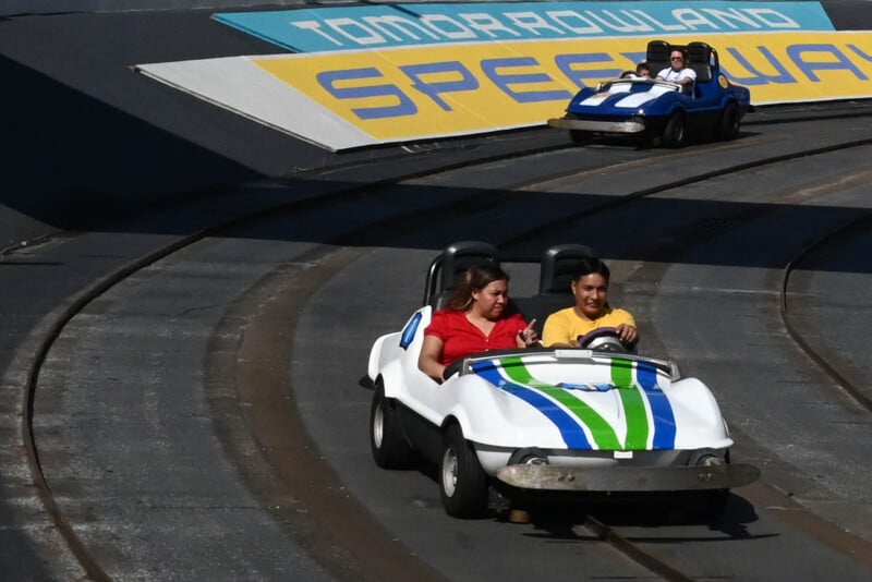 Two people drive a small white car with blue and green stripes on the Tomorrowland Speedway track. Another car with a driver and passenger follows. A large sign overhead reads "Tomorrowland Speedway.