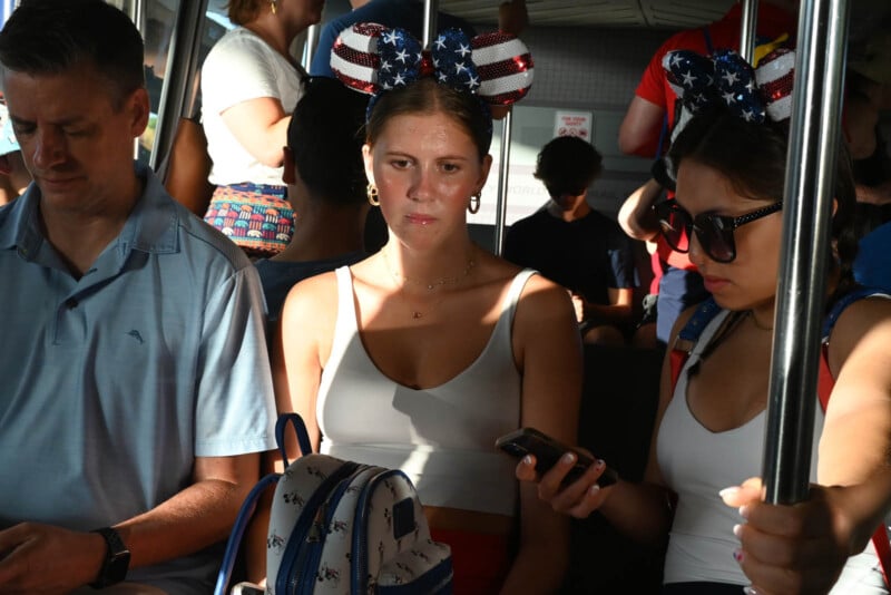 Three people are seated on public transit. A woman in the center wears patriotic Minnie Mouse ears and looks ahead, while another woman checks her phone. A man sits to the left, also looking at his phone. The sunlight casts shadows inside.