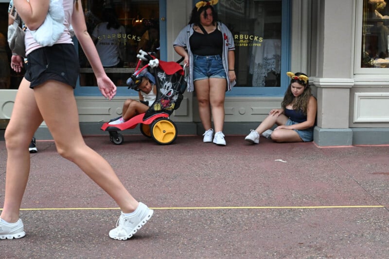 Three people are near a storefront: one sitting on the ground, one standing with arms crossed, and a child in a tricycle. Another person walks past wearing white sneakers. All wear Disney-themed headbands.