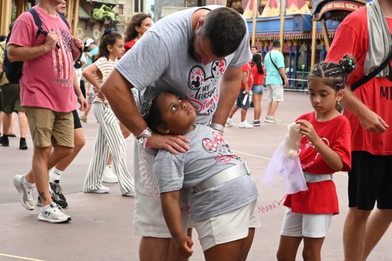 A man comforts a tired child wearing a Minnie Mouse shirt. Another child stands nearby, holding a toy. People walk in the background in a theme park setting, with colorful decorations.