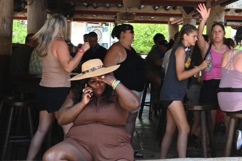 A woman sits on a low wall, talking on her cellphone and wearing a straw hat. Around her, several people are socializing and moving about inside a covered patio area with greenery visible in the background.