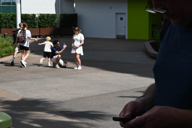 A group of people walk on a sunny day. A child runs ahead while two adults take a playful selfie. Another person is in the foreground, looking at a phone. Greenery and a building are visible in the background.