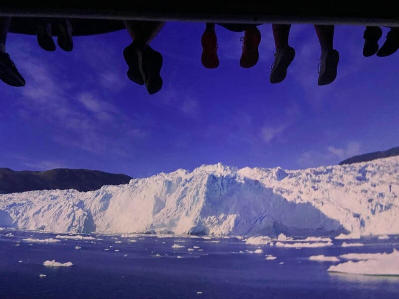 Feet hanging over the edge of a boat or ledge are silhouetted against a vibrant blue sky. Below, a vast ice shelf with white glaciers stretches across the horizon, with icy waters and floating ice chunks in the foreground.