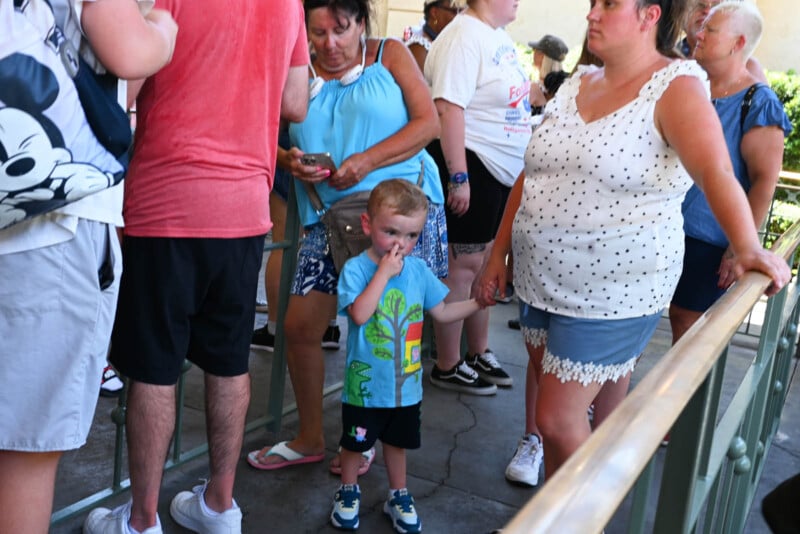 A young child in a colorful shirt stands in line, holding the hand of a woman in a white polka dot top. Several people are around them, some wearing themed clothing. The scene appears to be a busy outdoor queue.